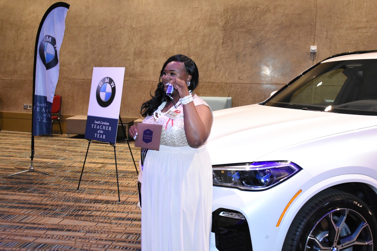 Chanda Jefferson, 2019-2020 South Carolina Teacher of the Year, holds up keys and stands next to her new wheels for the next year- a Mineral White Metallic BMW X5. 